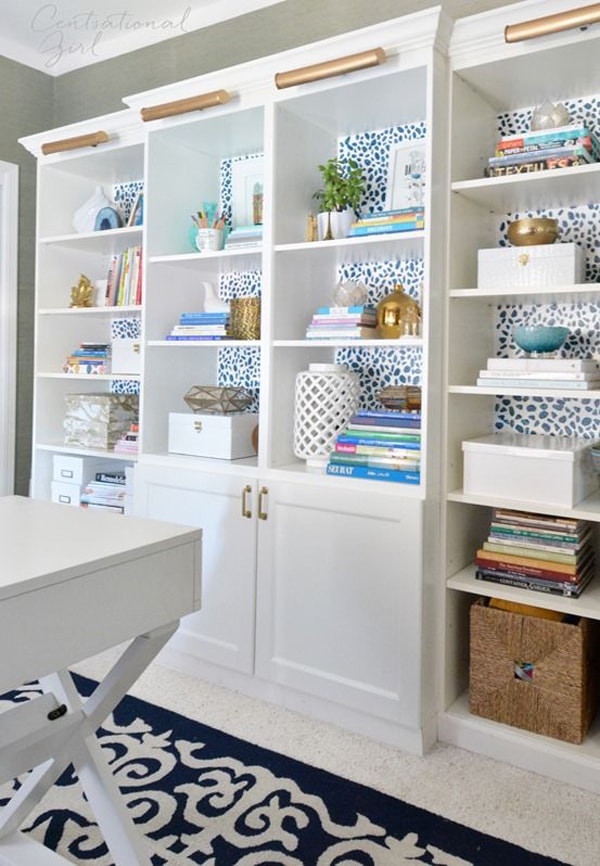 A white wooden built-in bookcase with a blue polka-dotted wallpaper background styled with accessories like books, storage containers, vaese, pictures and artwork