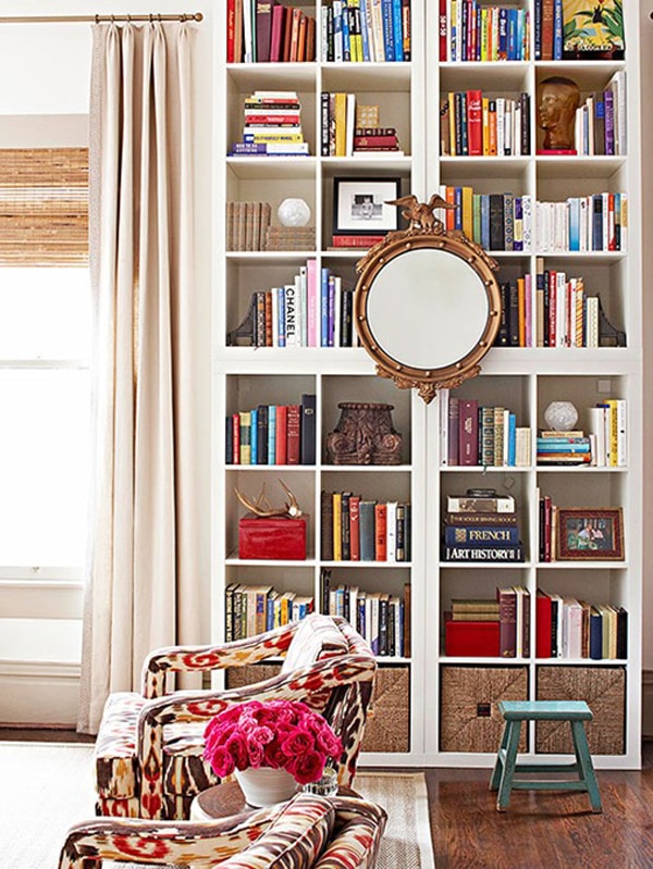 A white wooden bookcase with cube shelves, styled with a mirror in the center and filled with colorful stacks of books