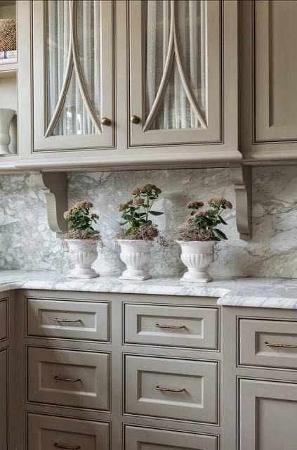 Beige colored cabinets with marble counter top decorated with 3 white urns with flowers.
