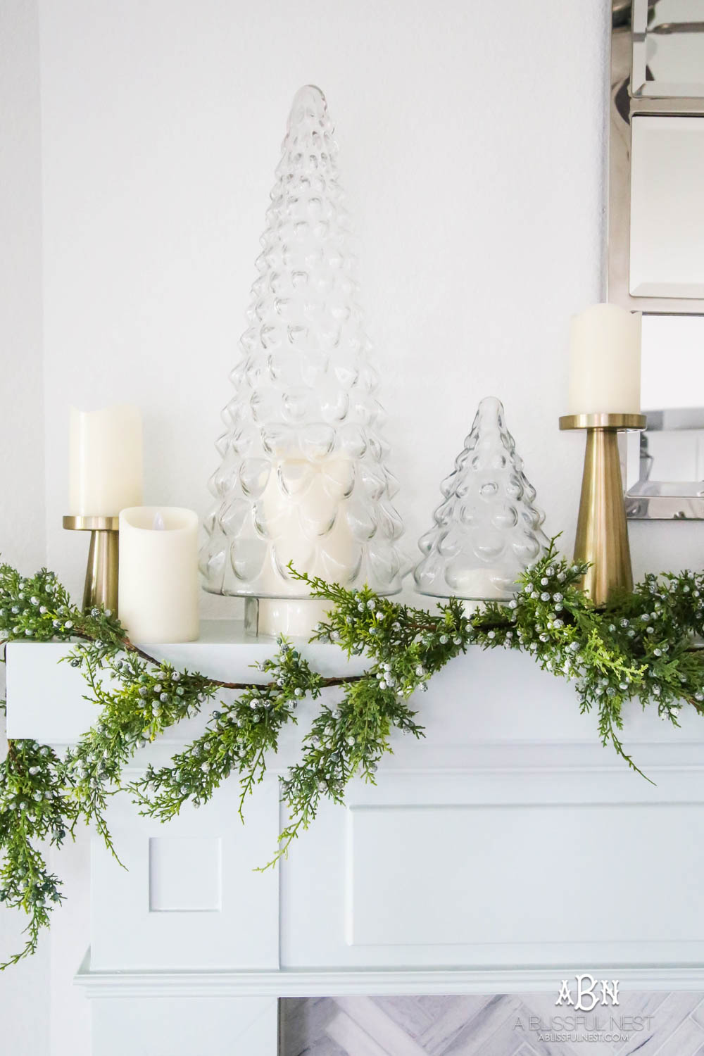 Gorgeous blue and silver Christmas mantle décor with juniper berry garland and glass hurricane Christmas trees. Simple and festive holiday décor in this open concept living space. Check out all the white, silver and gold Christmas decor in this holiday home tour on ablisfulnest.com. #ABlissfulNest #Christmasdecor #Christmasdecorating #CoastalChristmasdecor #christmastree #christmasmantle