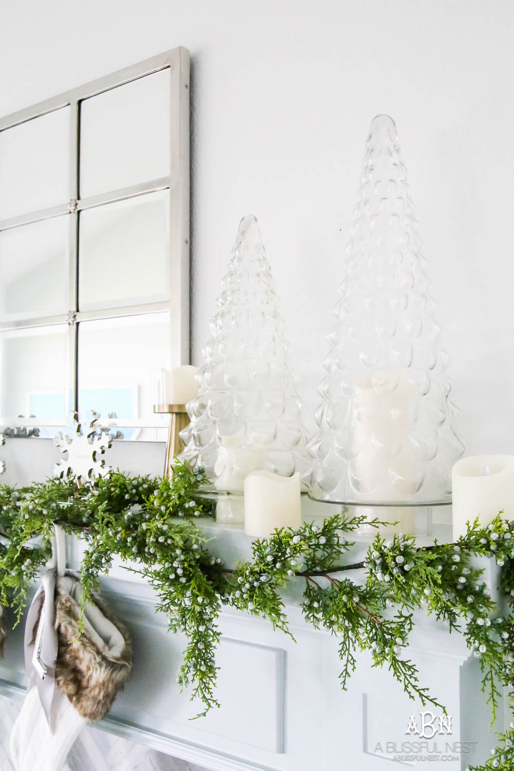 Gorgeous blue and silver Christmas mantle décor with juniper berry garland and glass hurricane Christmas trees. Simple and festive holiday décor in this open concept living space. Check out all the white, silver and gold Christmas decor in this holiday home tour on ablisfulnest.com. #ABlissfulNest #Christmasdecor #Christmasdecorating #CoastalChristmasdecor #christmastree #christmasmantle