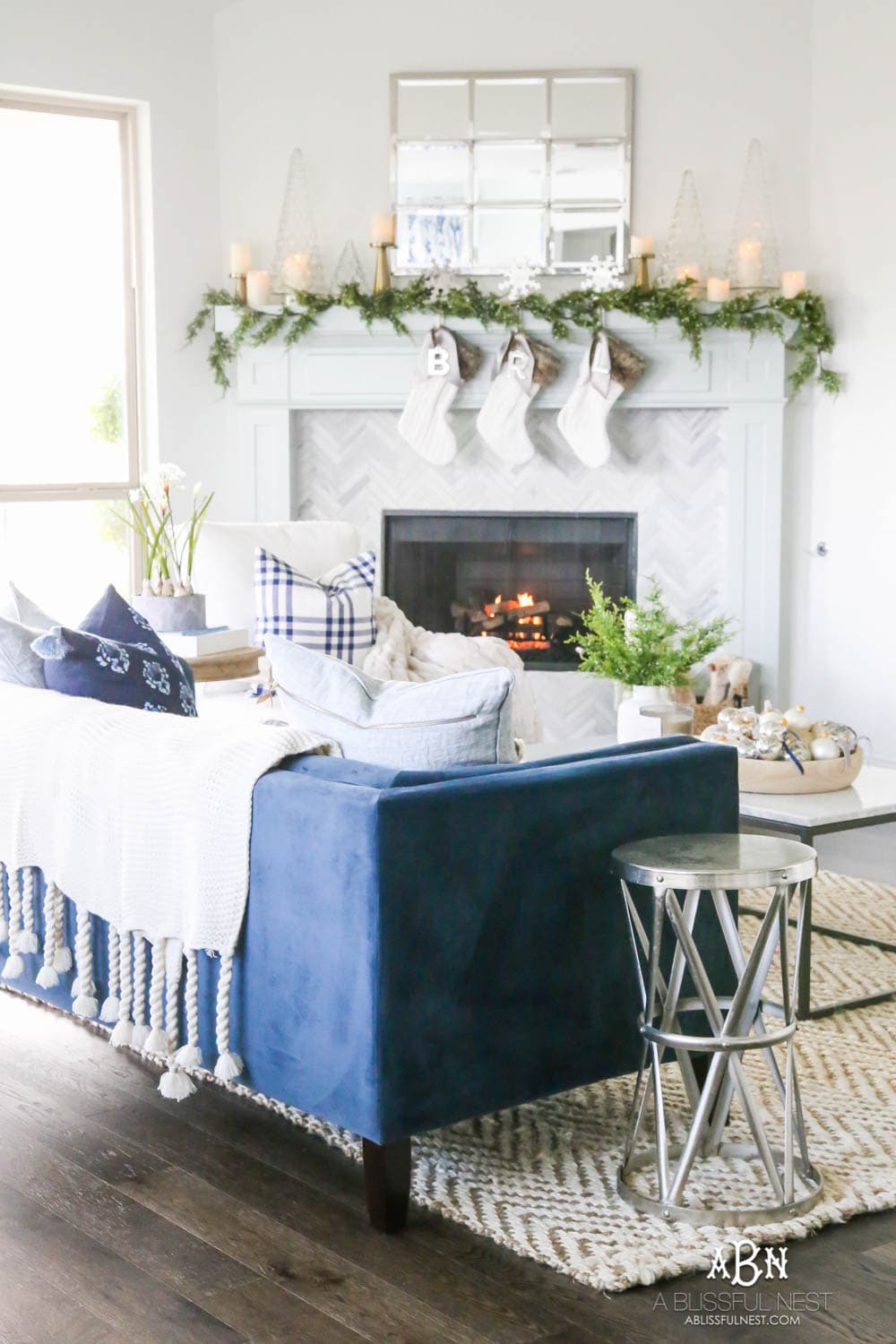 Gorgeous blue and silver Christmas mantle décor with juniper berry garland and glass hurricane Christmas trees. Simple and festive holiday décor in this open concept living space. Check out all the white, silver and gold Christmas decor in this holiday home tour on ablisfulnest.com. #ABlissfulNest #Christmasdecor #Christmasdecorating #CoastalChristmasdecor #christmastree #christmasmantle