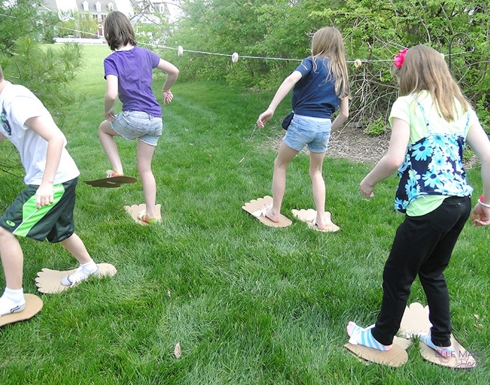 kids walking through the grass wearing giant cardboard foot cutouts on their feet