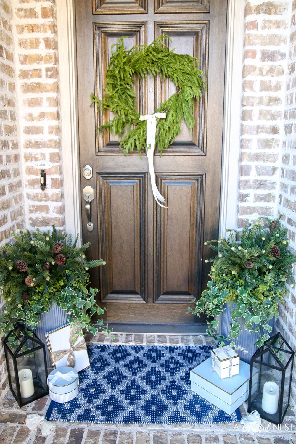 Brick entryway with solid wood door, pine wreath with a white satin ribbon, blue and white doormat with planters and black lanterns with gift boxes tucked next to them.