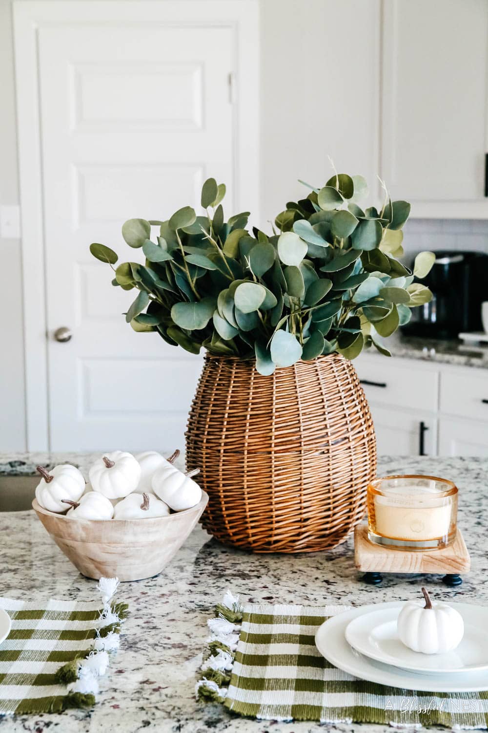 Woven basket filled with eucalyptus leaves with a small bowl of white pumpkins and a fall candle sitting on a kitchen counter.
