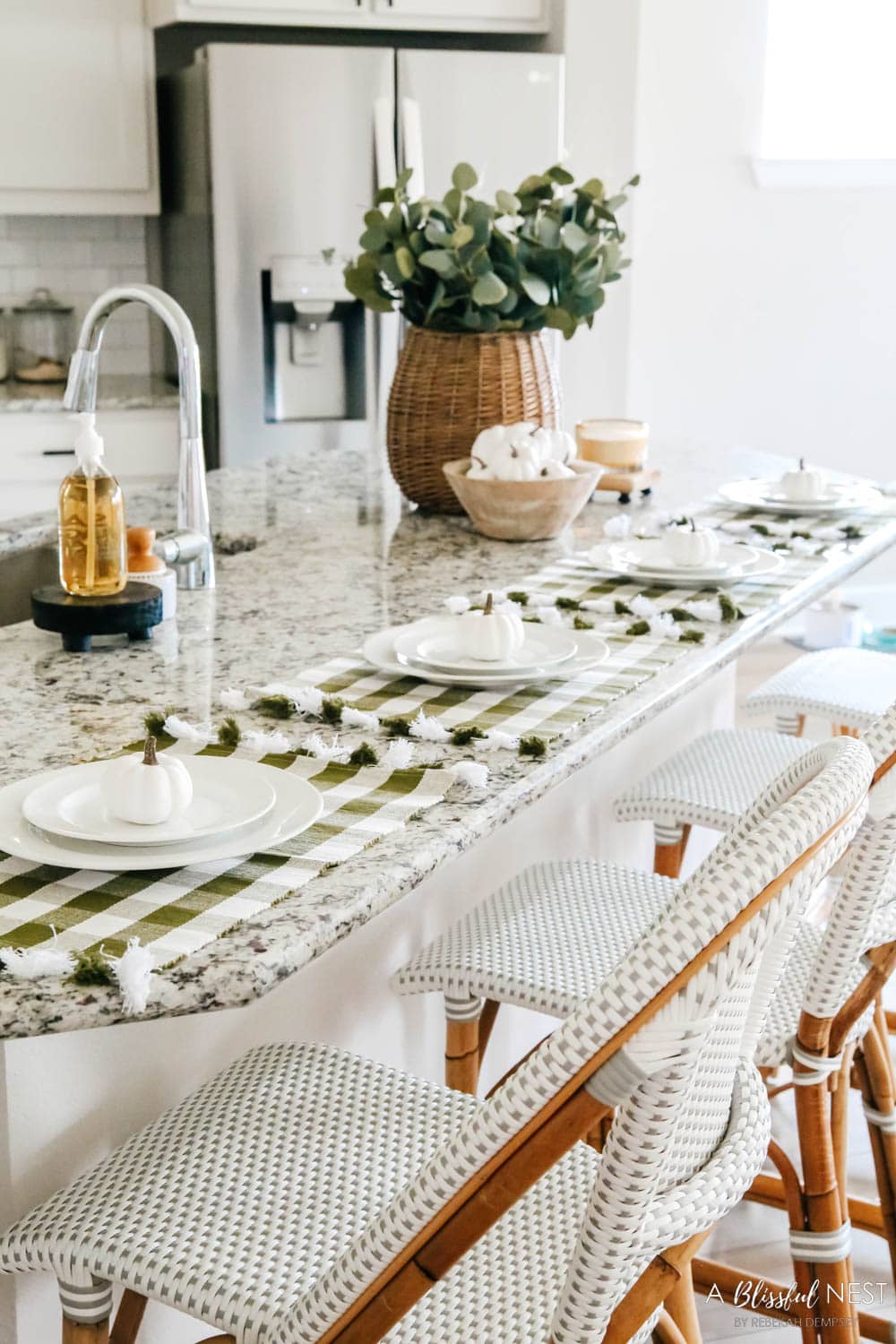 Green and white cloth placemats with white plates and small mini white pumpkins on a breakfast counter in a kitchen.