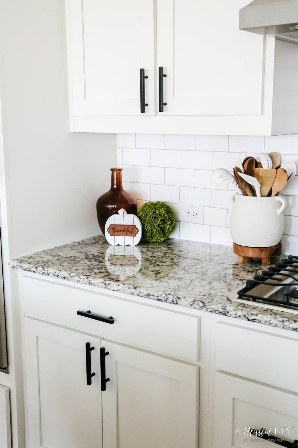 Fall decor clustered together on the corner of a kitchen counter, white pumpkin, amber vase, kitchen utensils in a white canister.