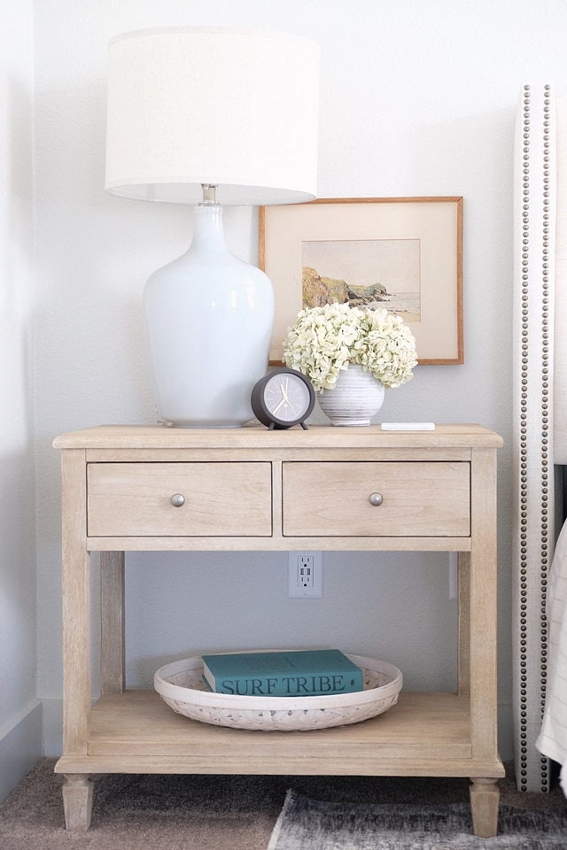 Light wood nightstand with shelf and drawer, white table lamp, small art on wall.