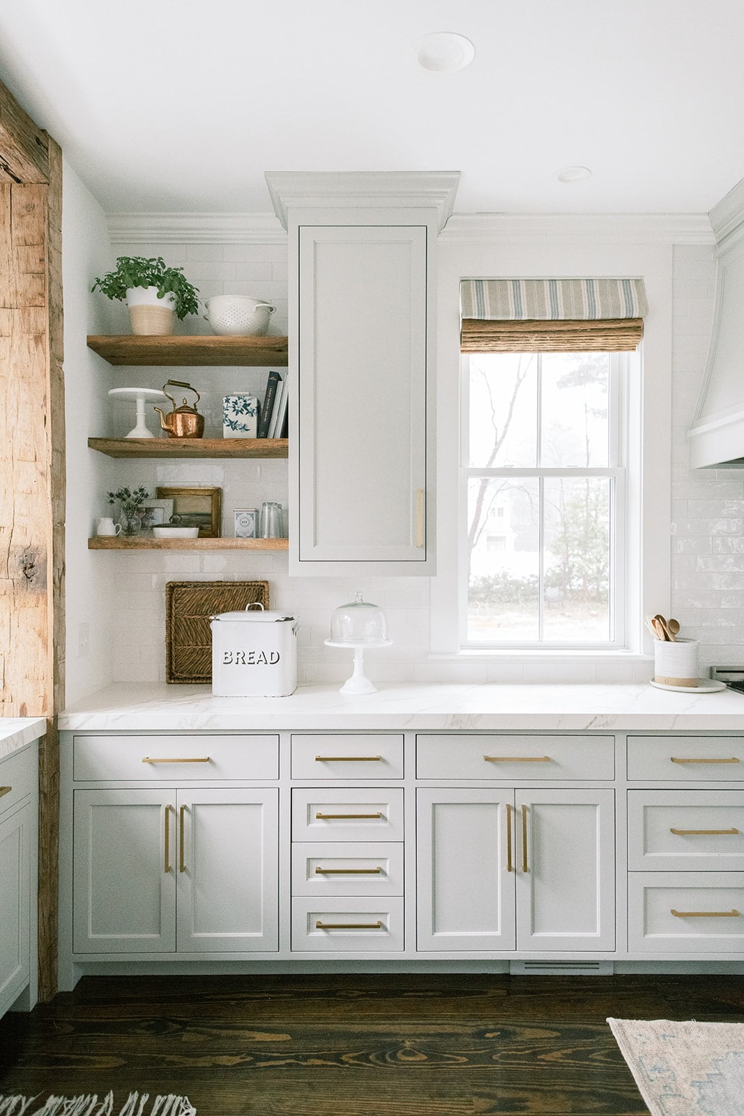 Light grey kitchen cabinets with gold pulls and wood floating shelves. White subway tile and quartz countertops.