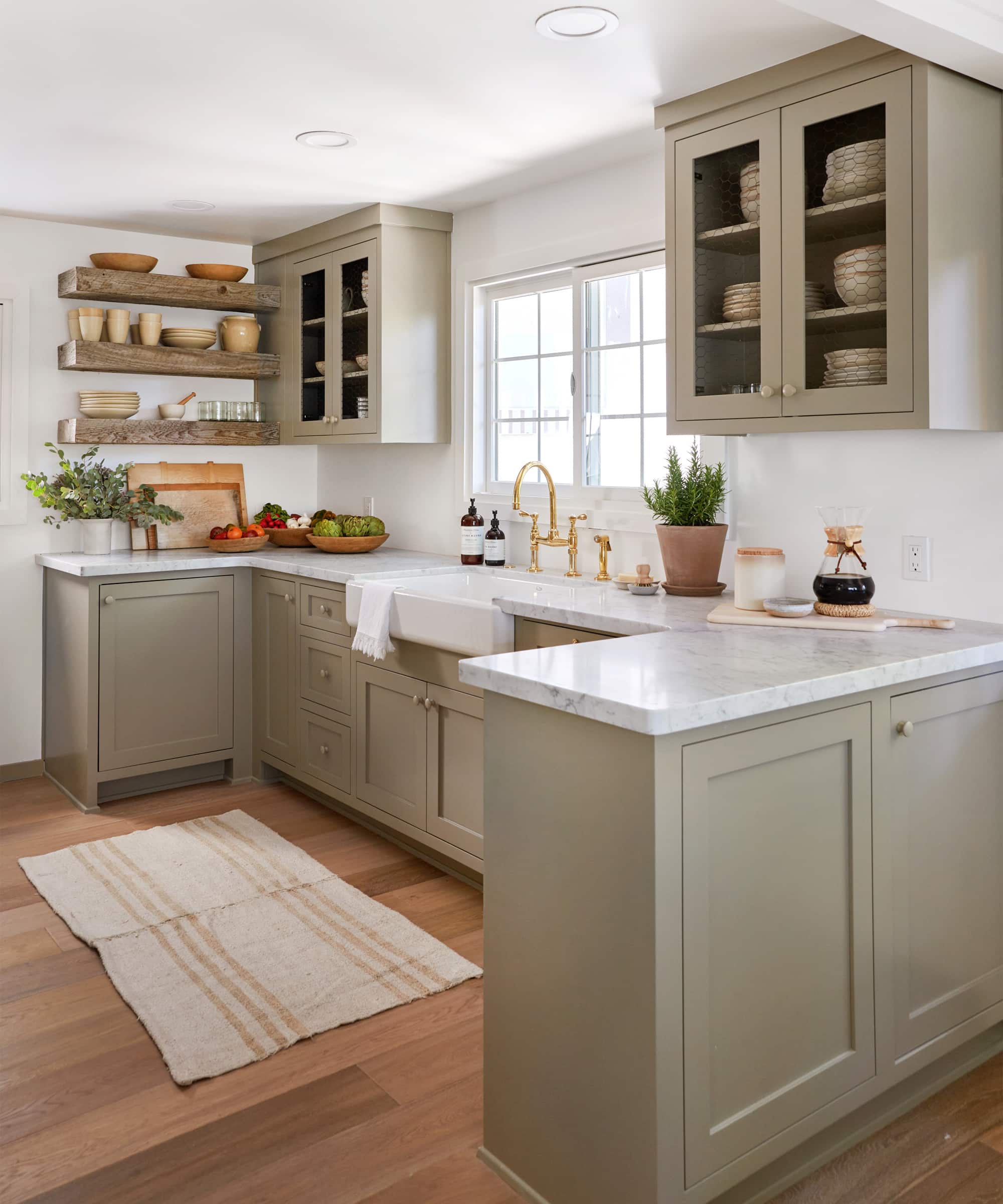 greige cabinets, gold faucet, marble countertops, wood floating shelves, window over sink, glass cabinets