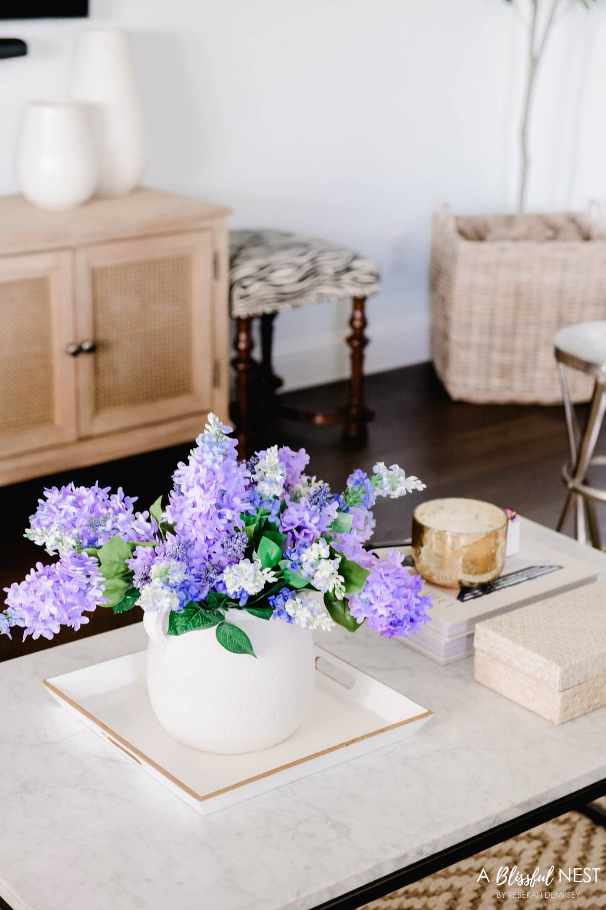 white vase sitting on a white tray with a gold candle and rattan box holding remotes for the tv.