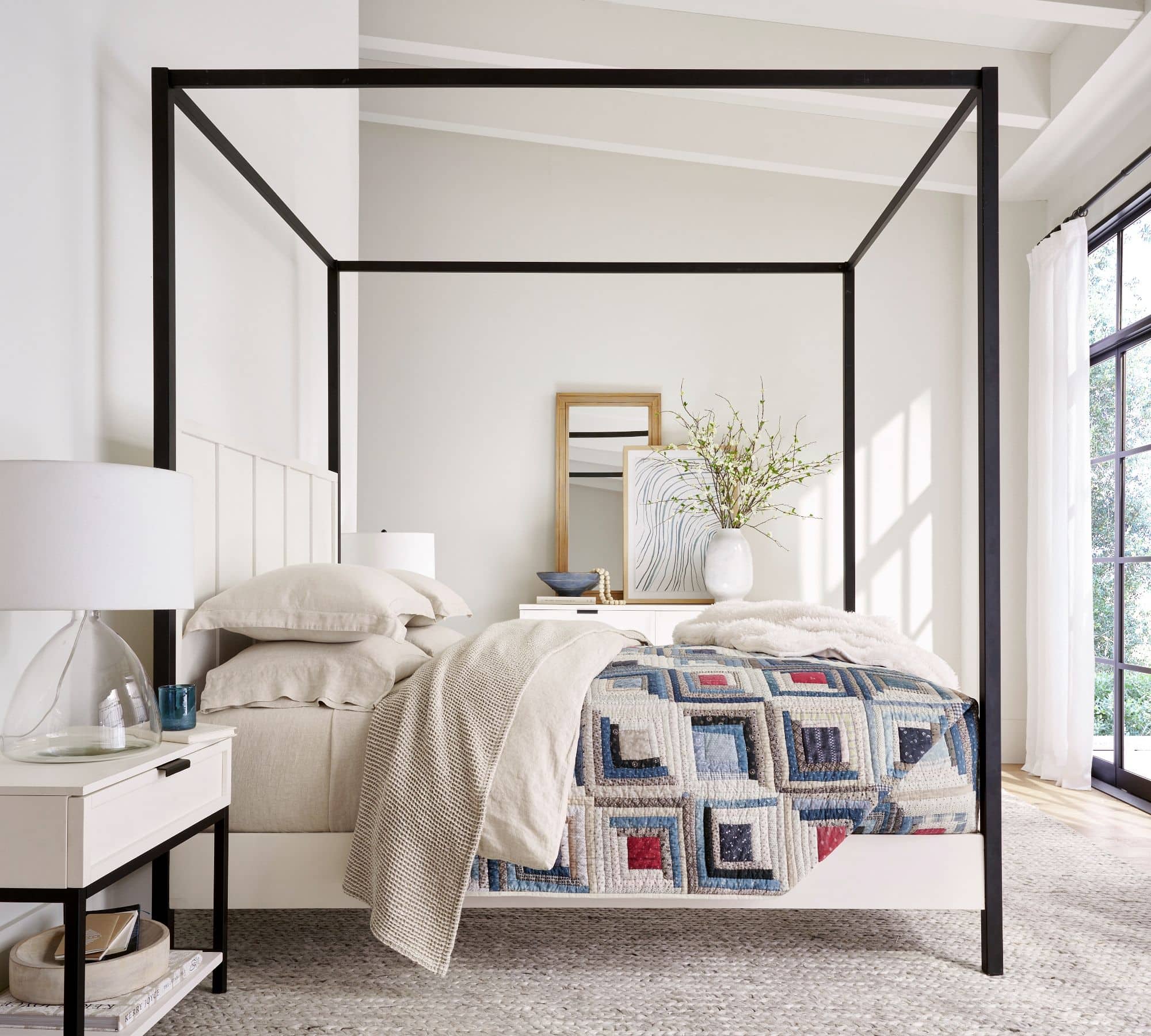 Black framed bed with cream colored bedding, patterned quilt and beige knit blanket at end of bed