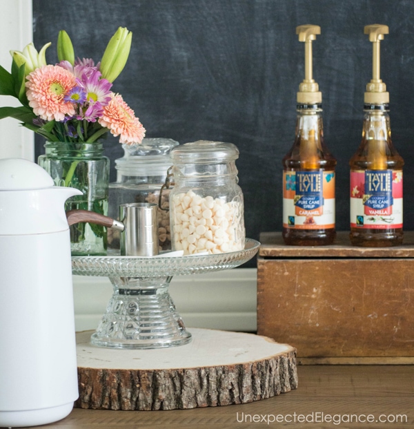 glass cake stand with mason jars with marshmallows in it. Cake stand sitting on a lice of wood. Coffee syrups next to it.