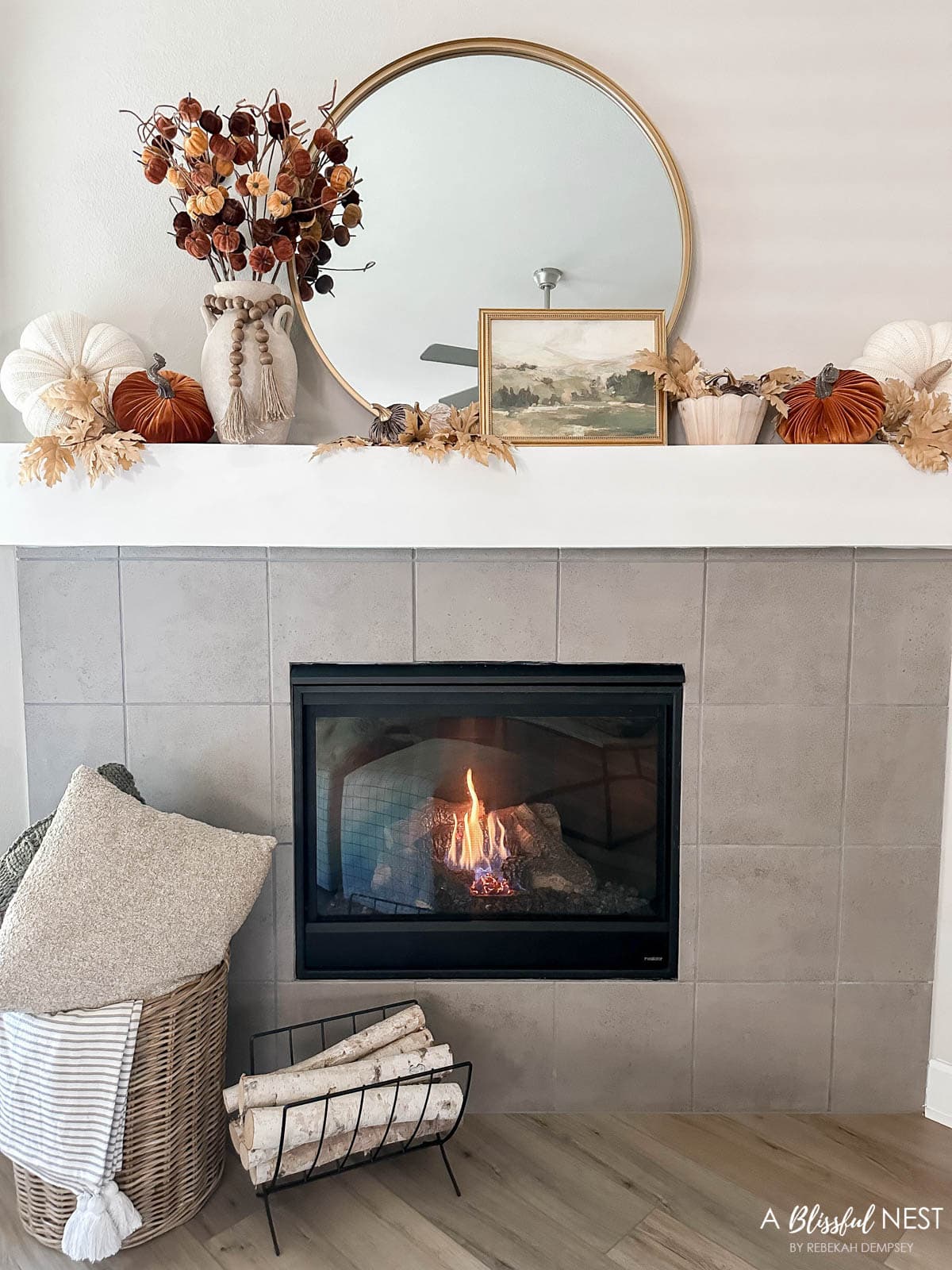 fall mantel with pumpkins, gold round mirror, fall leaf picks and a fireplace burning. 