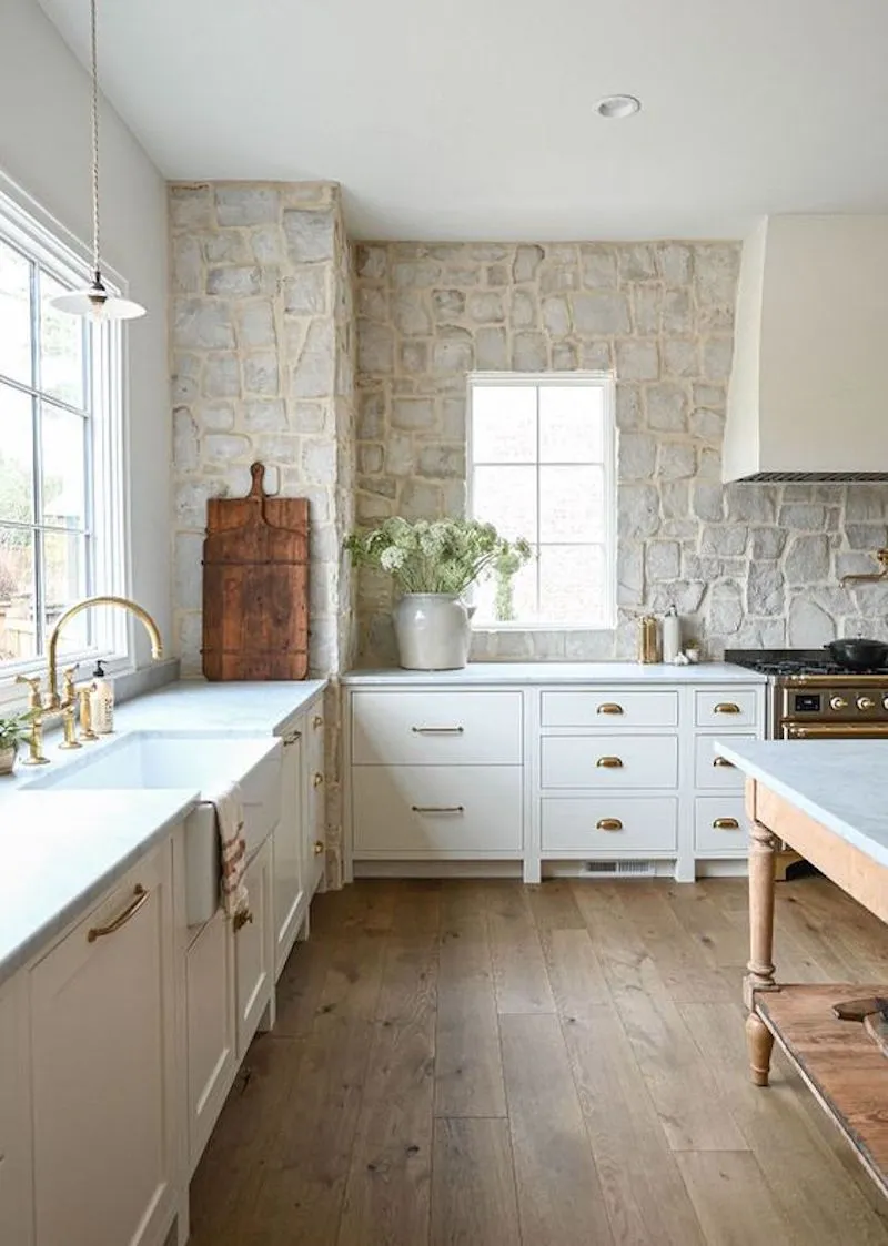 Stone walls, white cabinets, gold cabinet hardware, light wood flooring in this kitchen design