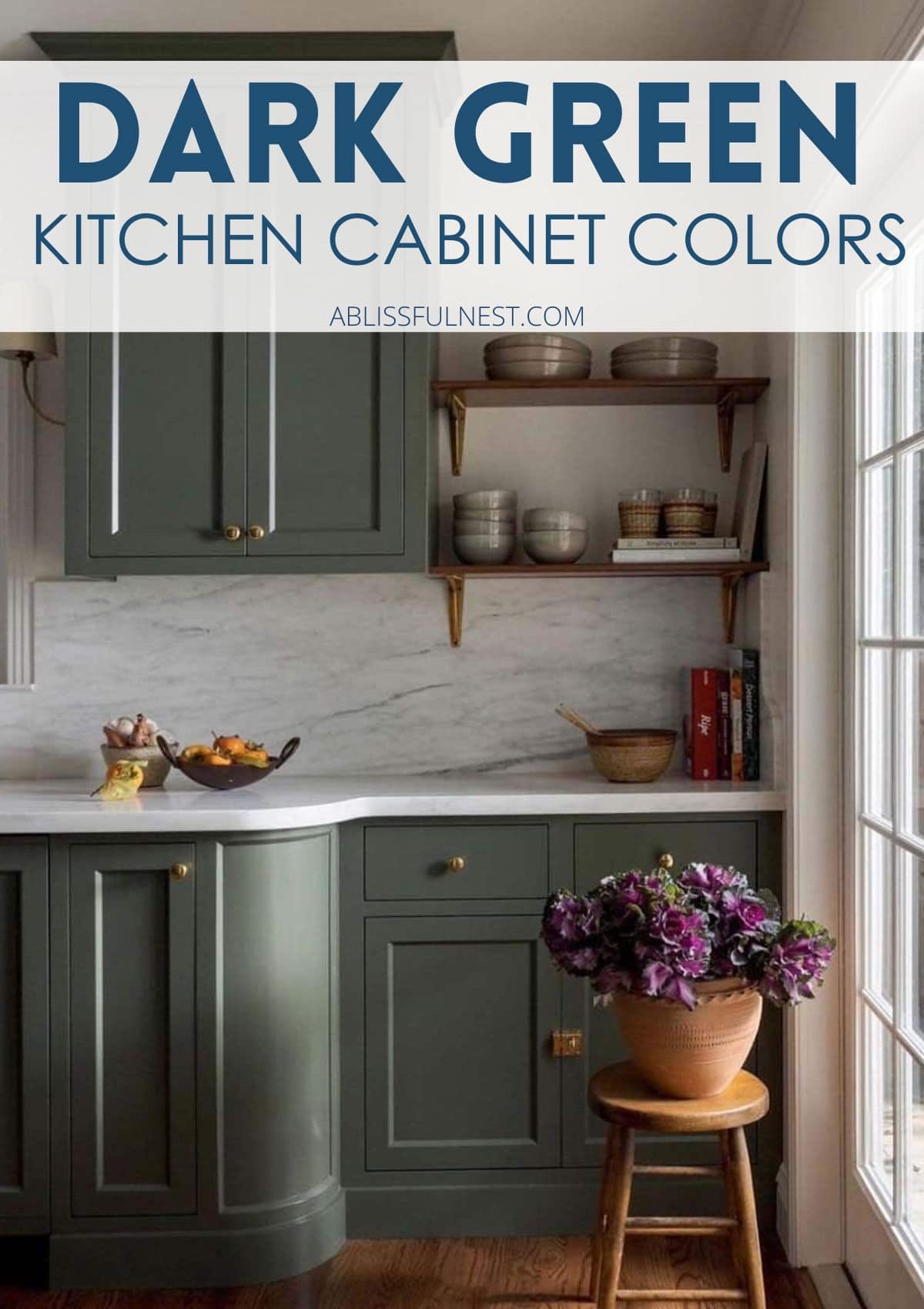 Dark green kitchen cabinets with gold hardware and a wood stool with a garden pot filled with purple flowers. 